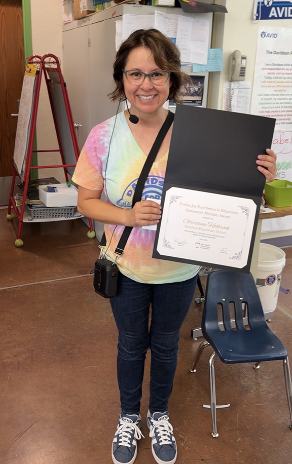 A woman in a tie-dye shirt proudly holds up her award certificate